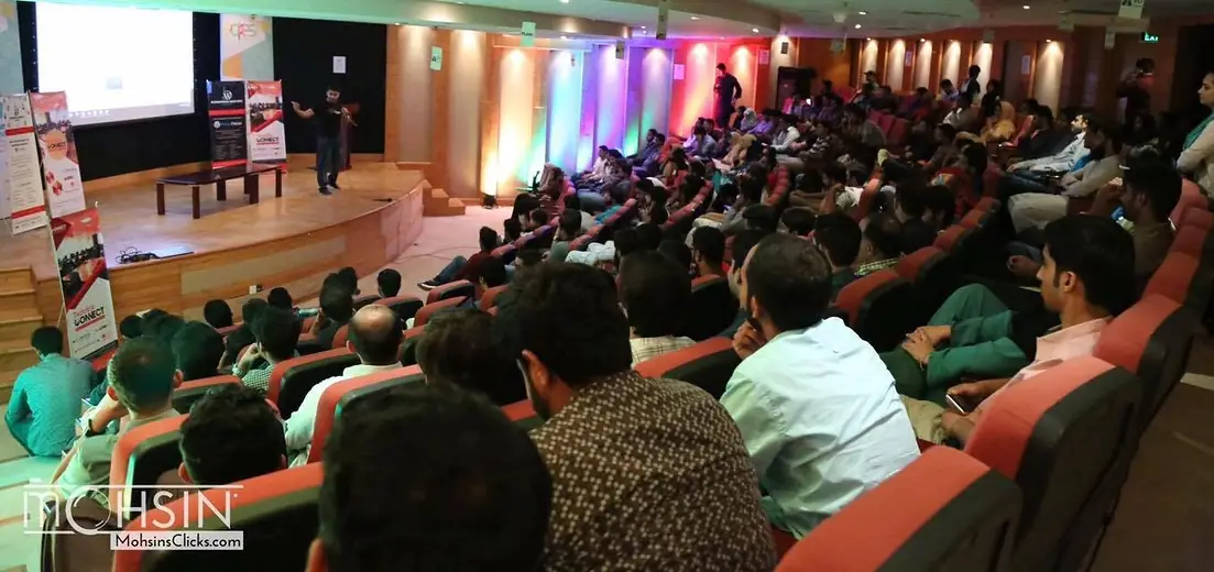 Audience in a tiered auditorium facing a speaker on stage